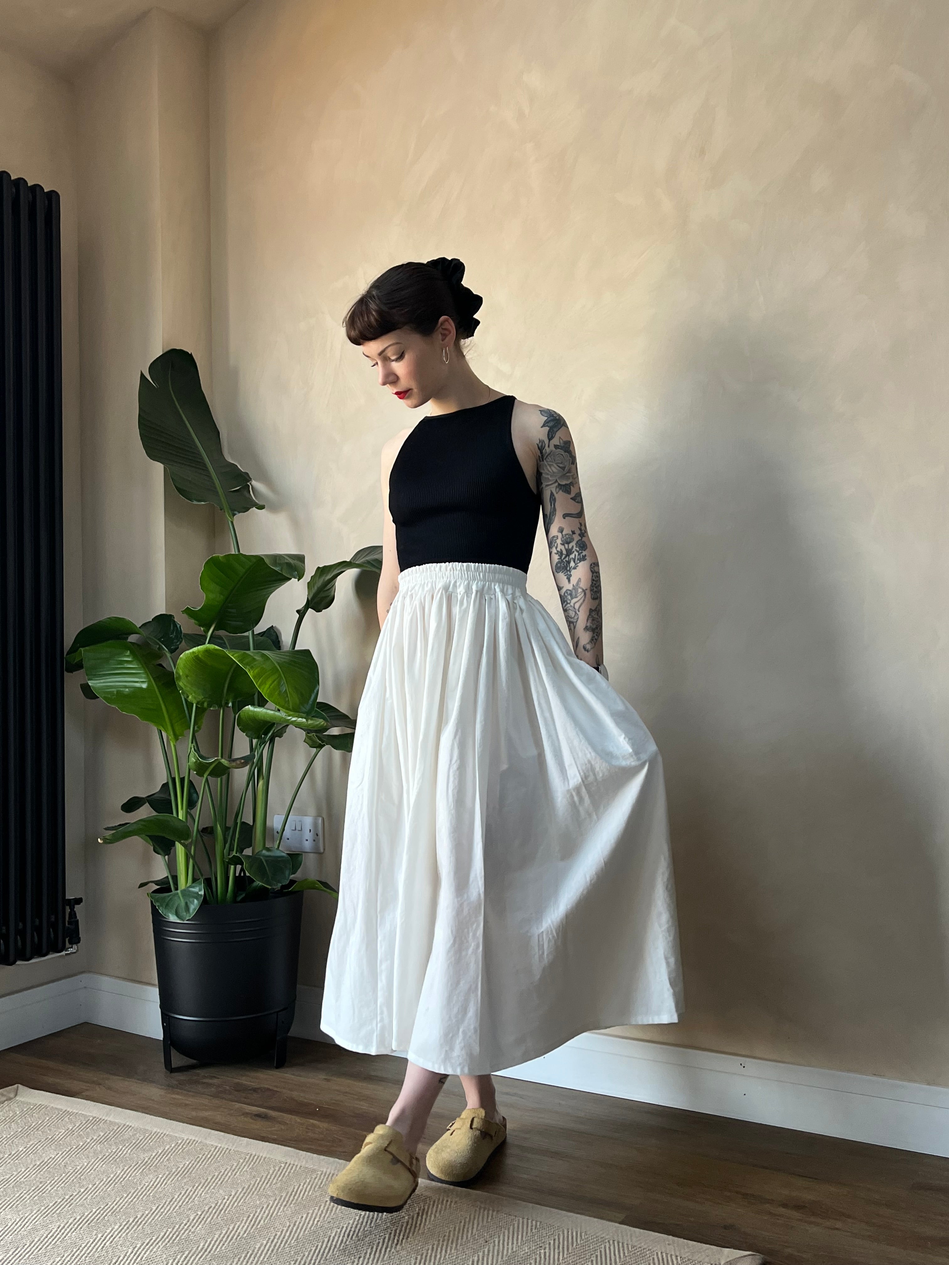 A hand holding a midi skirt in white, with a plant in the background. The skirt appears to be made from sustainable cotton fabric.