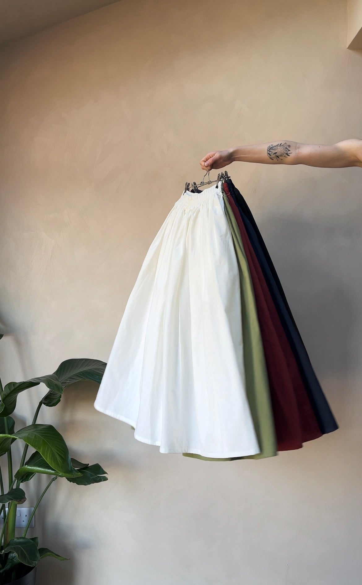 A hand holding a midi skirt in white, with a plant in the background. The skirt appears to be made from sustainable cotton fabric.