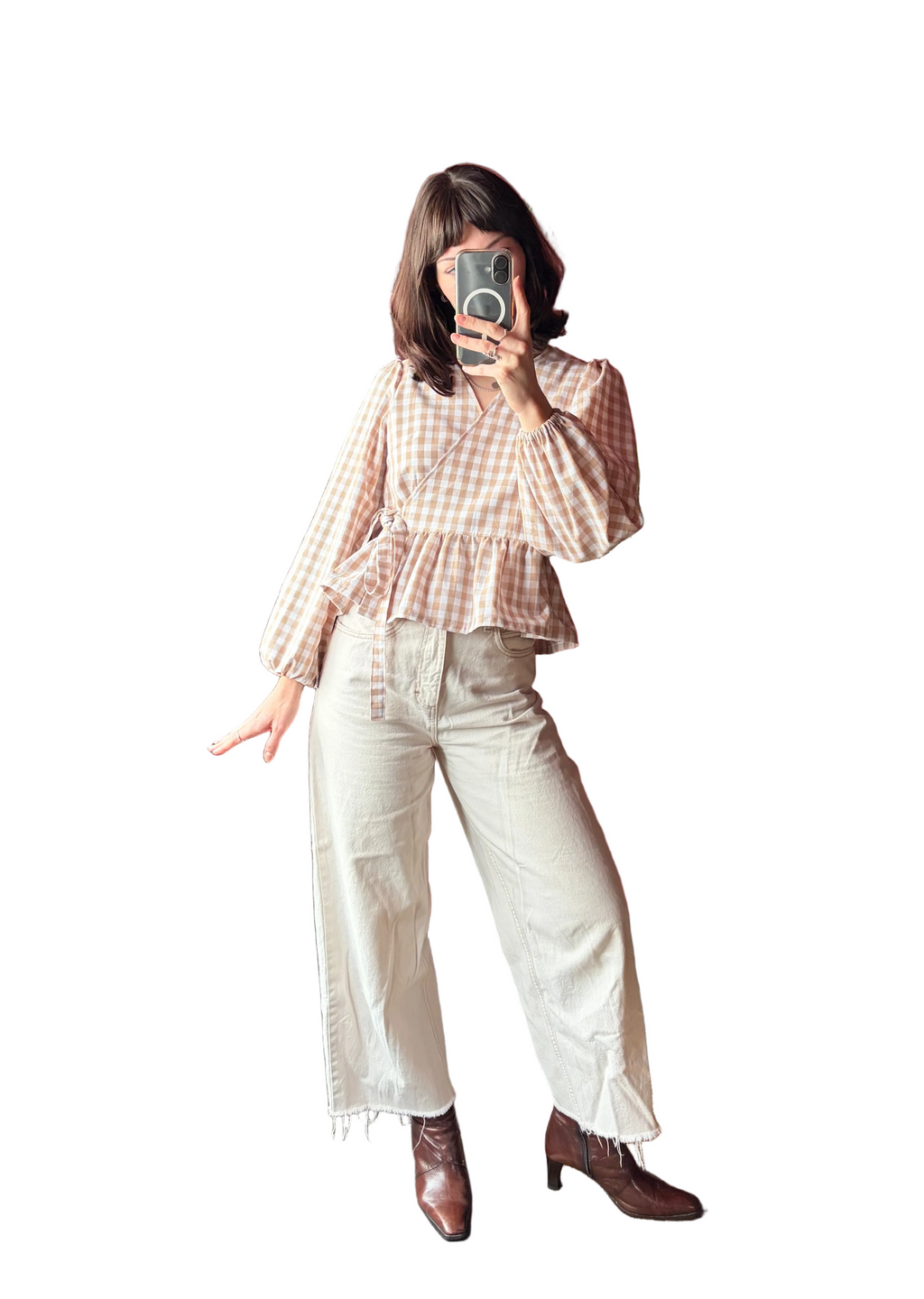 A woman takes a mirror selfie in a bedroom showing an outfit consisting of a beige and white gingham long-sleeve peplum top, cream wide-leg cropped jeans, and brown leather ankle boots. The background features a dark red wall with built-in shelves and plants.