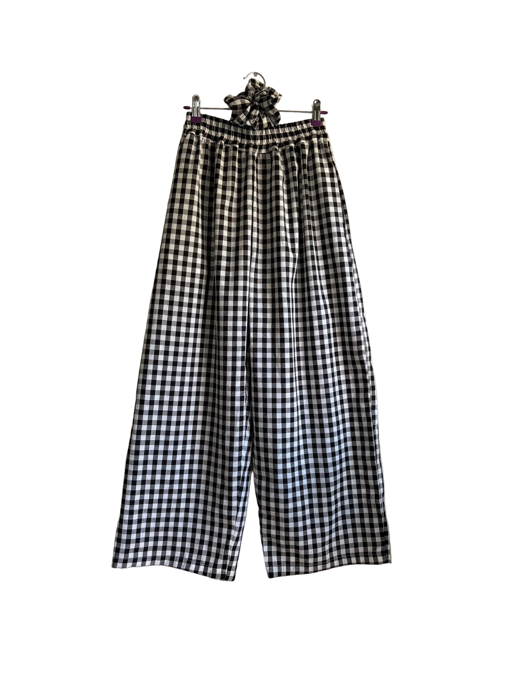 Black and white gingham wide-leg cropped pants with an elastic waistband and a matching fabric scrunchie detail at the top, hanging on a wall next to a plant.