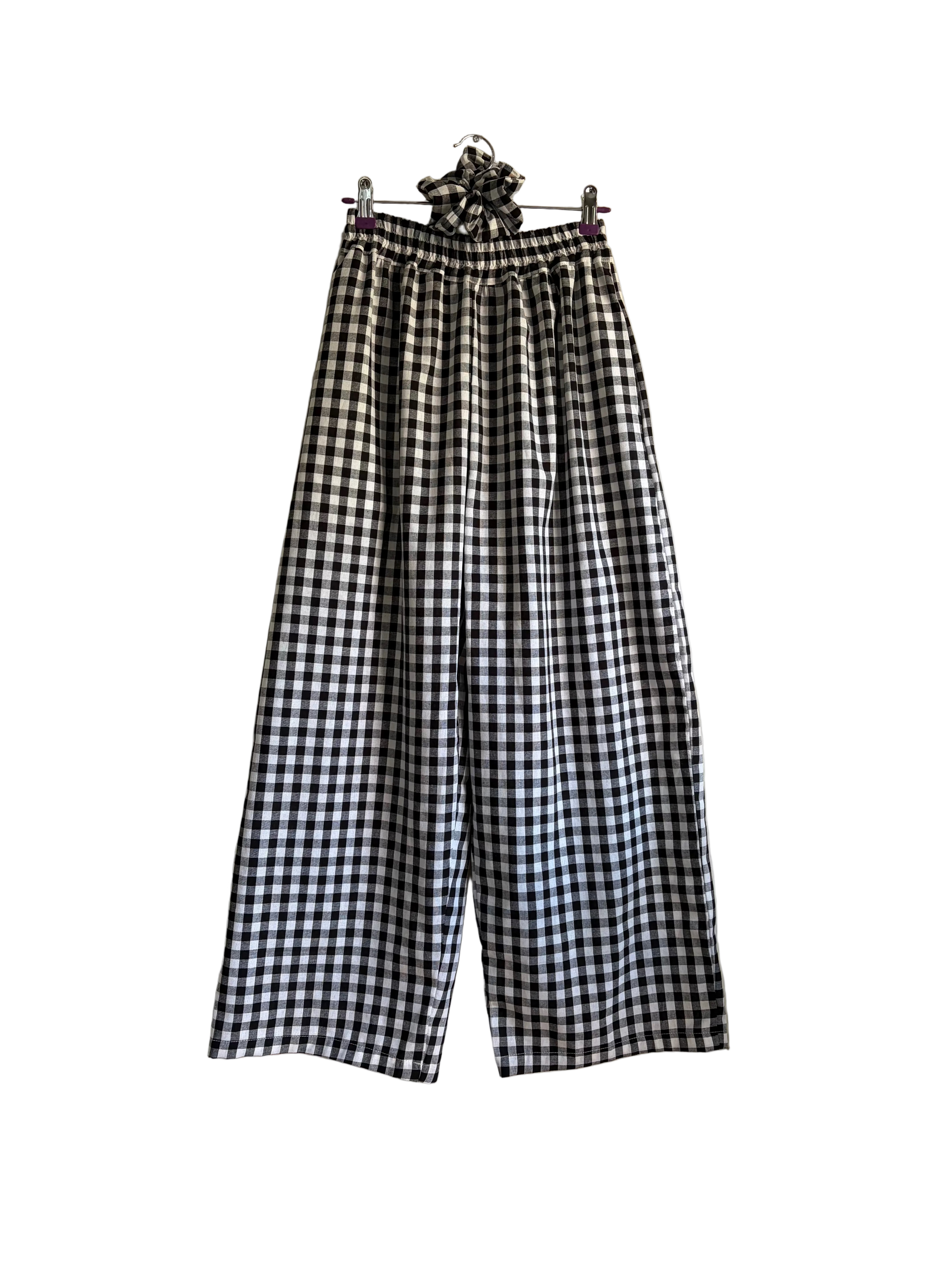 Black and white gingham wide-leg cropped pants with an elastic waistband and a matching fabric scrunchie detail at the top, hanging on a wall next to a plant.
