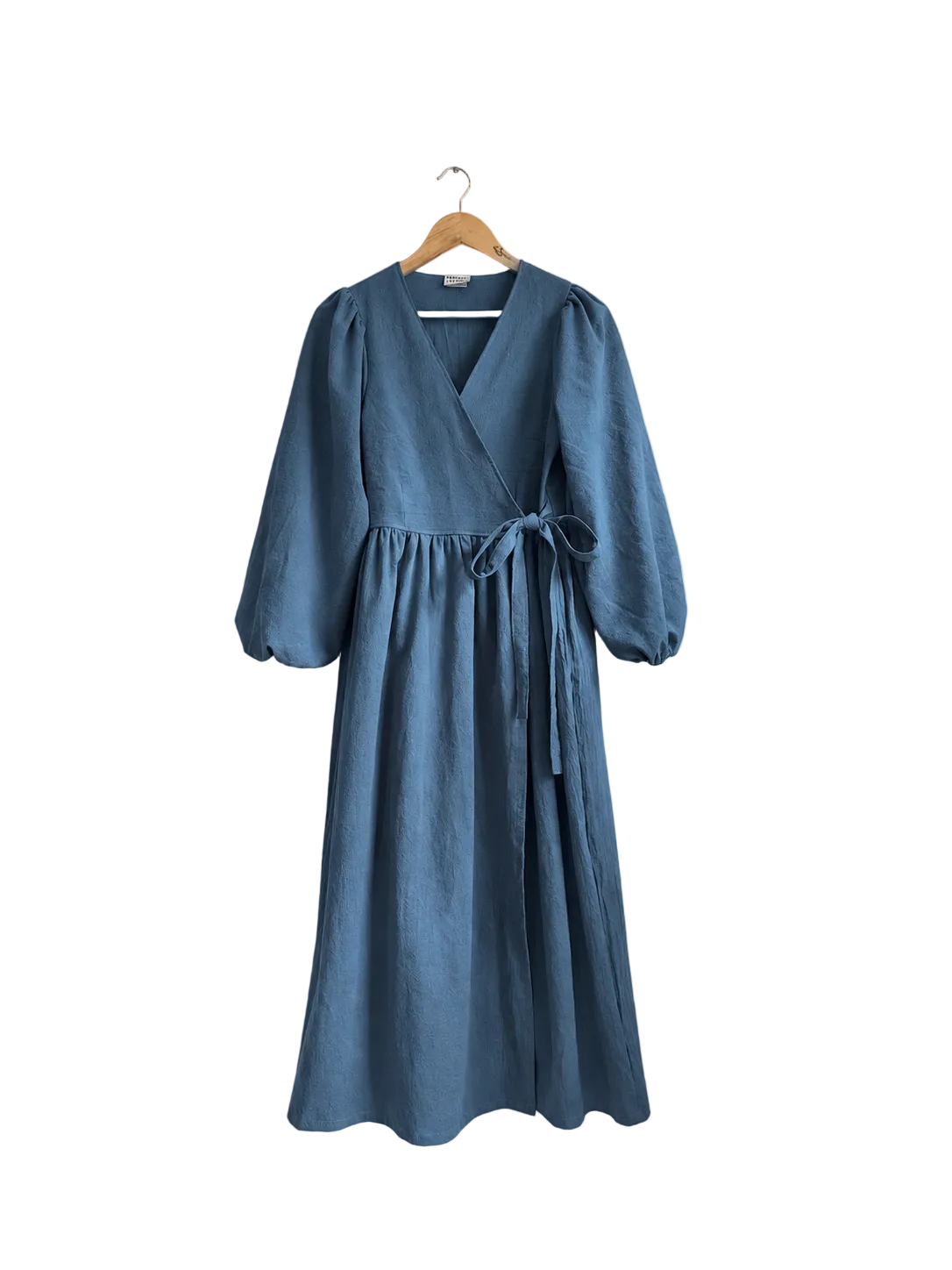 A dusty blue linen wrap dress with three-quarter sleeves and a tie waist, hanging on a wooden hanger against a beige wall next to a curtain and a green plant.