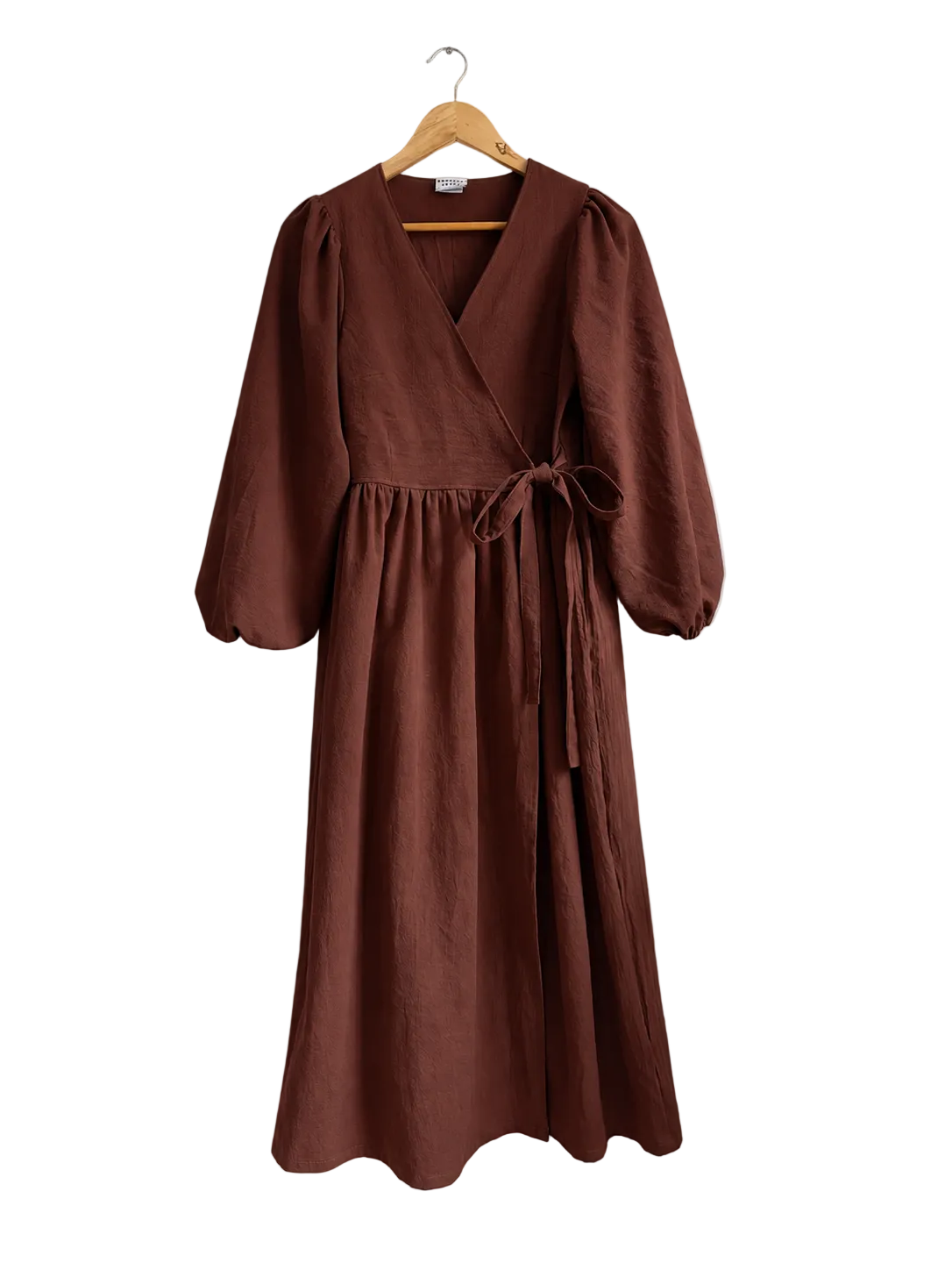 A brown linen wrap dress with three-quarter puff sleeves and a self-tie waist belt, hanging on a wooden hanger against a neutral off-white wall.