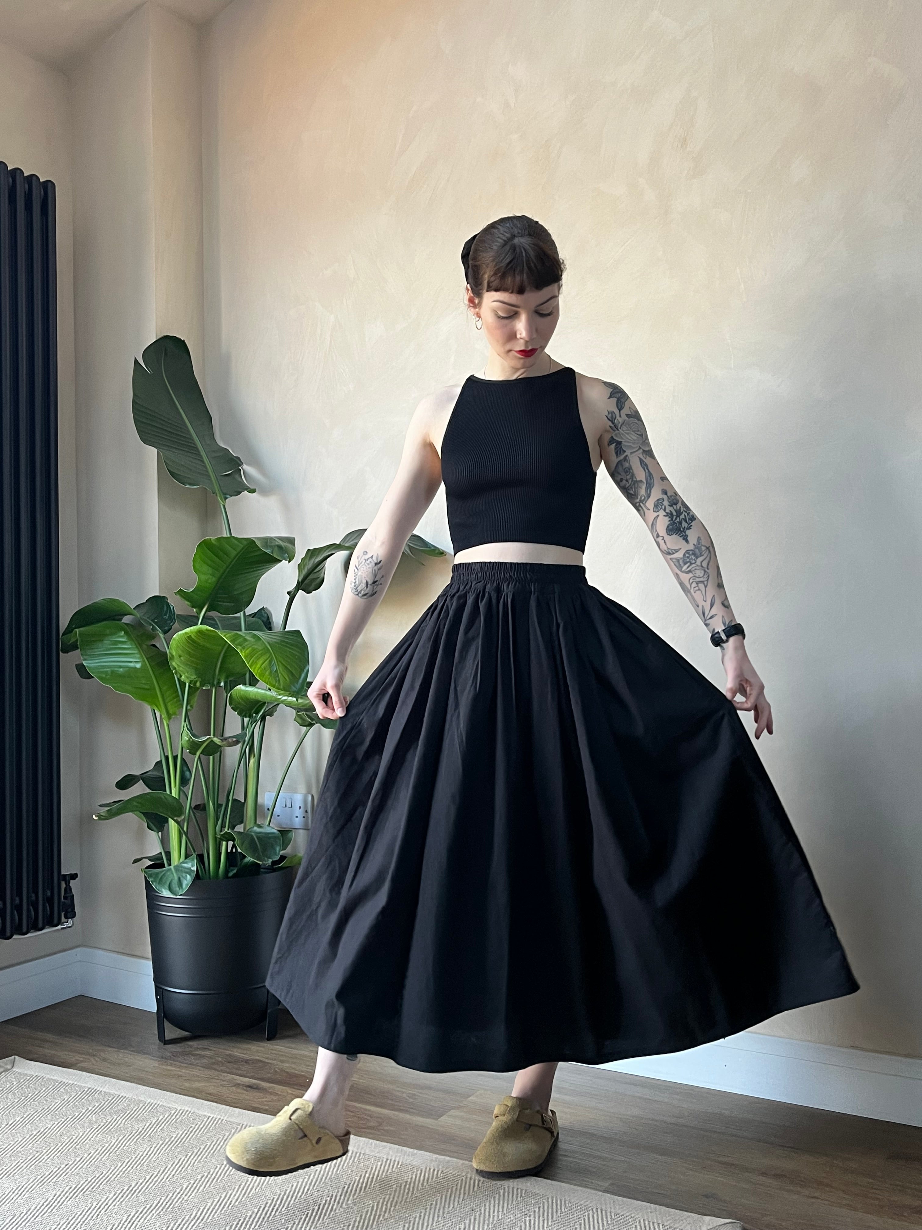 A hand holding a midi skirt in white, with a plant in the background. The skirt appears to be made from sustainable cotton fabric.