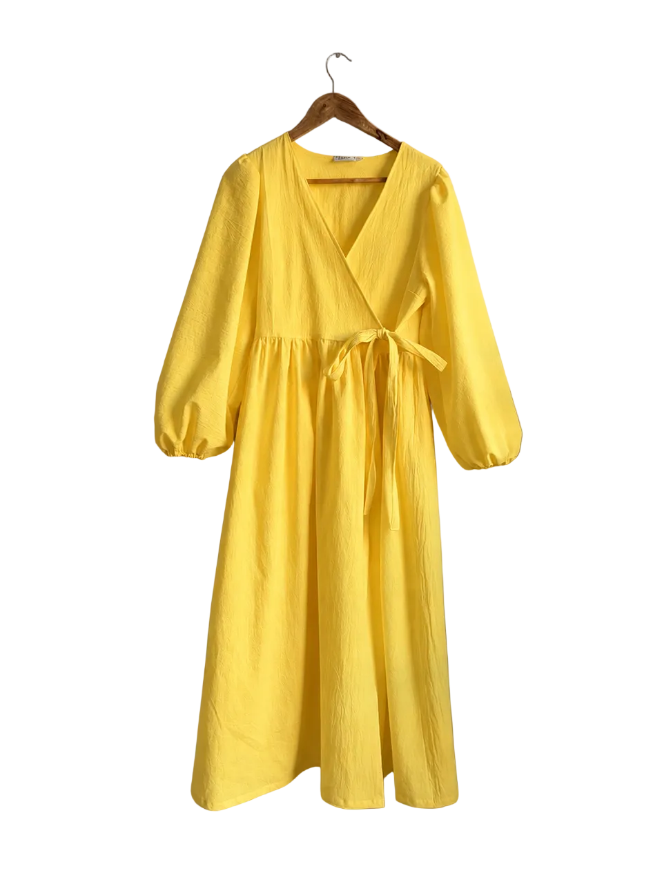 A bright yellow wrap-style midi dress with three-quarter puff sleeves hangs on a wooden hanger against a neutral beige wall, with a curtain and a potted plant visible on the right side.