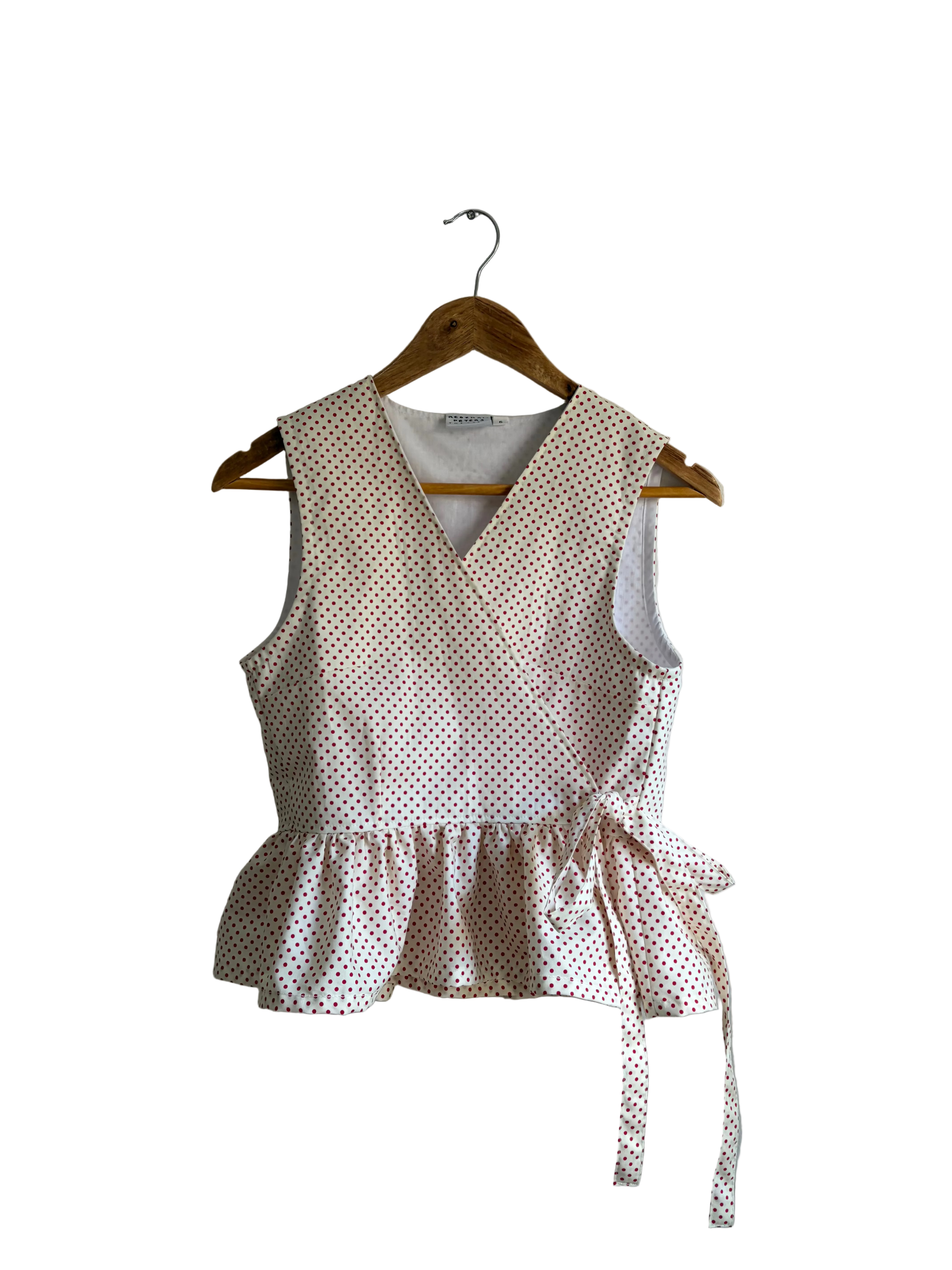 A sleeveless wrap-style top with a ruffled hem and side tie, featuring a small white polka dot pattern on a light pink background, hanging on a wooden hanger against a plain wall.