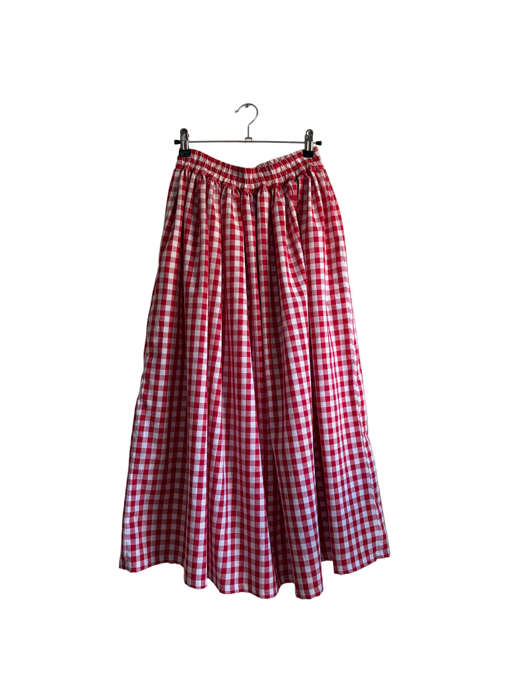 A red and white gingham pleated midi skirt hanging from a black clothes hanger against a beige wall, with a curtain and green plant leaves visible on the right side.