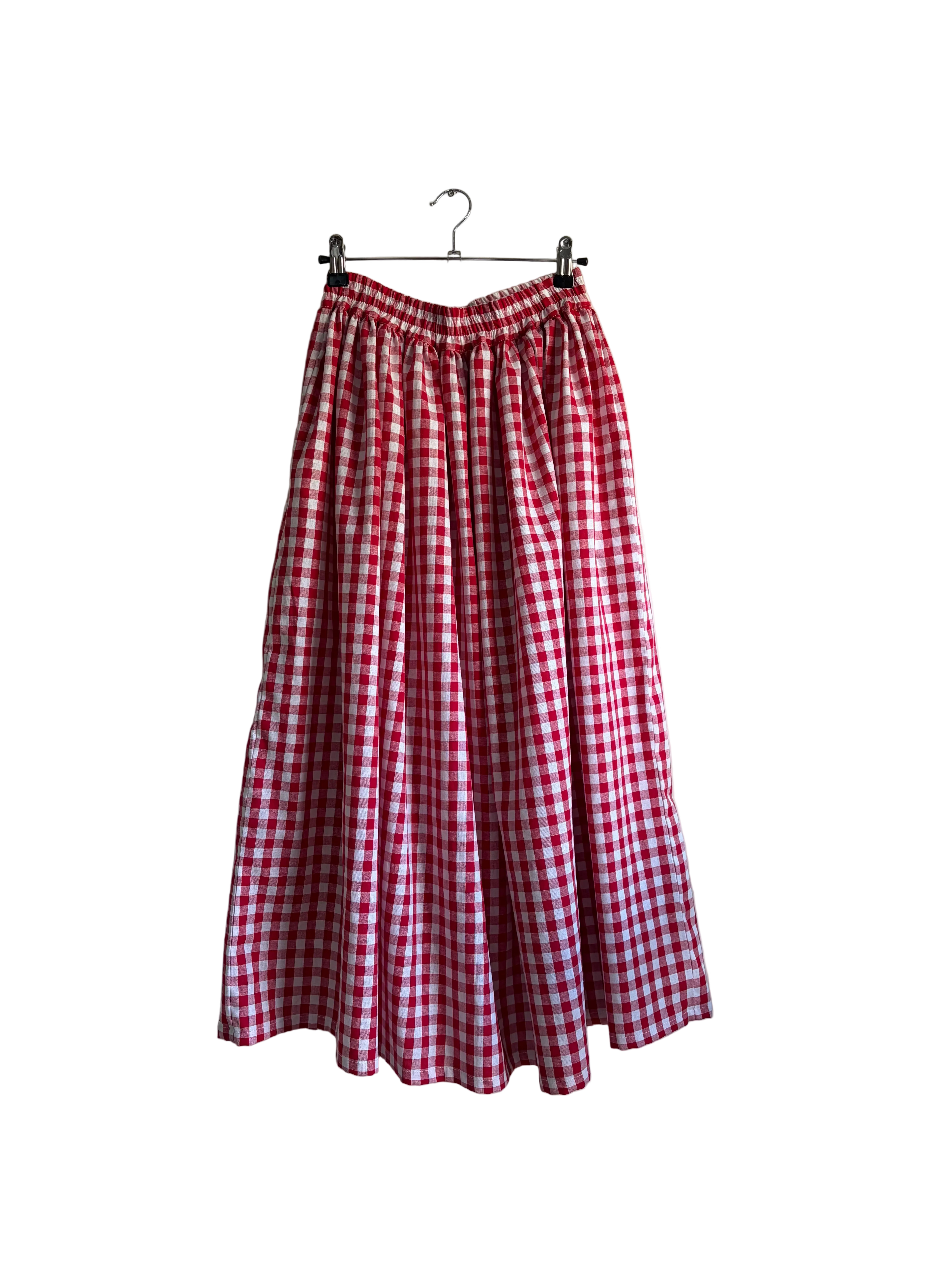 A red and white gingham pleated midi skirt hanging from a black clothes hanger against a beige wall, with a curtain and green plant leaves visible on the right side.