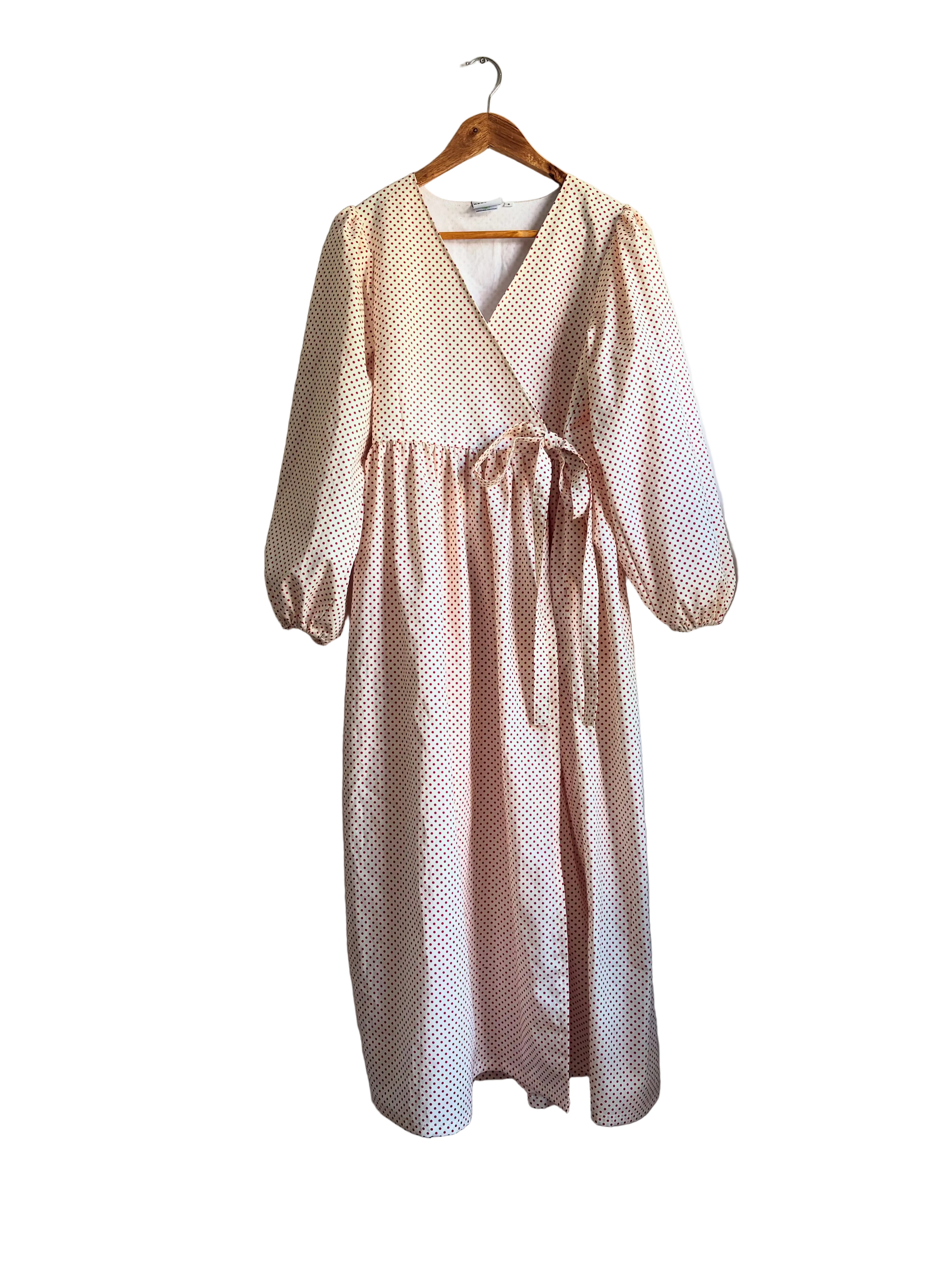 A light pink and white polka dot wrap dress with three-quarter sleeves hangs on a wooden hanger against a neutral wall next to a curtain and a plant.