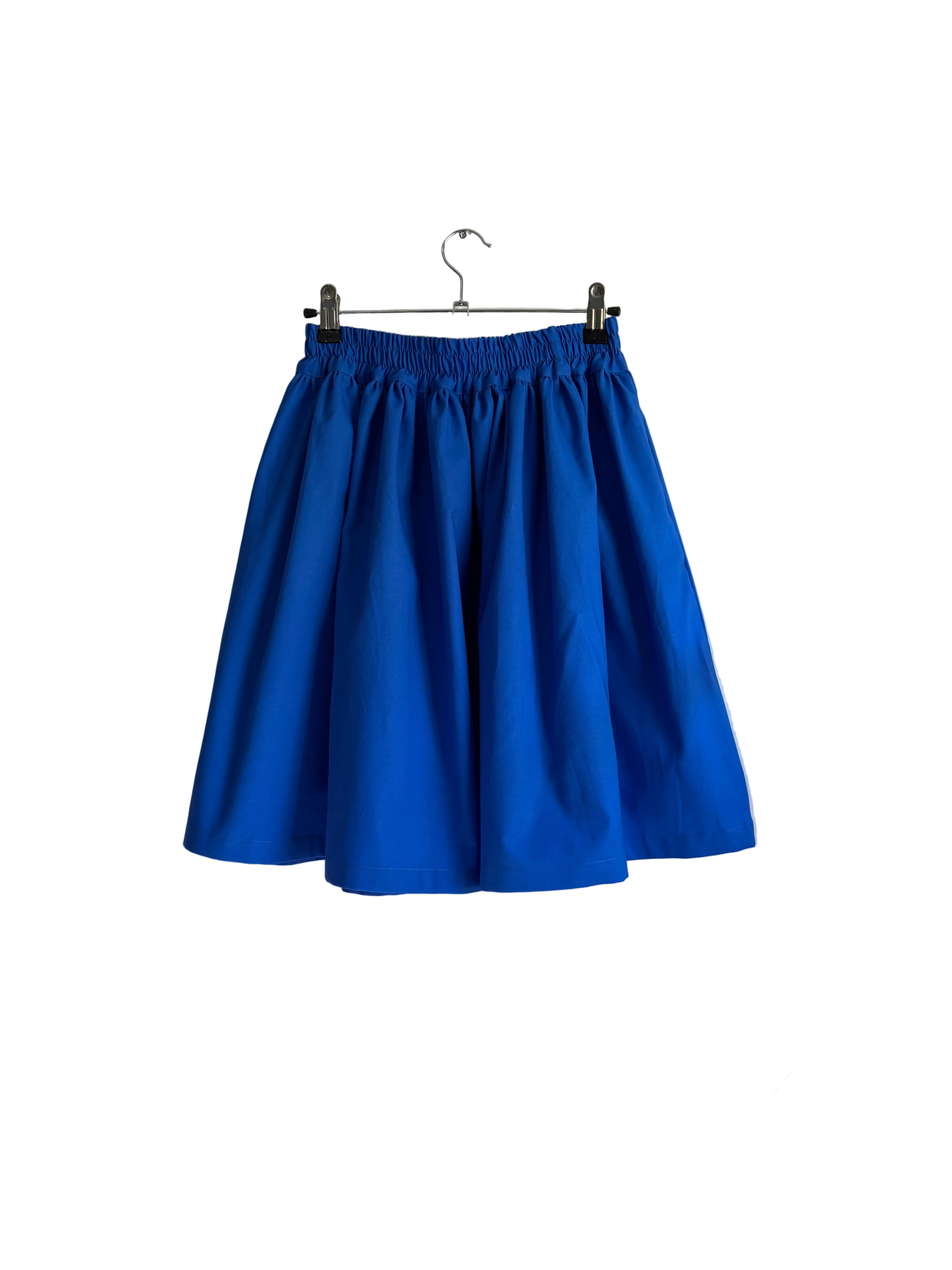 A bright blue pleated skirt hanging from a metal hanger against an off-white wall.