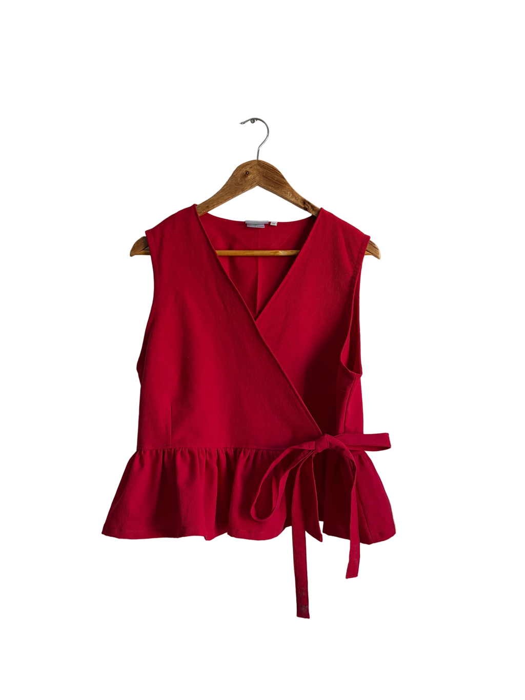 A bright red sleeveless wrap-style top with a ruffled hem and side tie, hanging on a wooden hanger against an off-white wall.