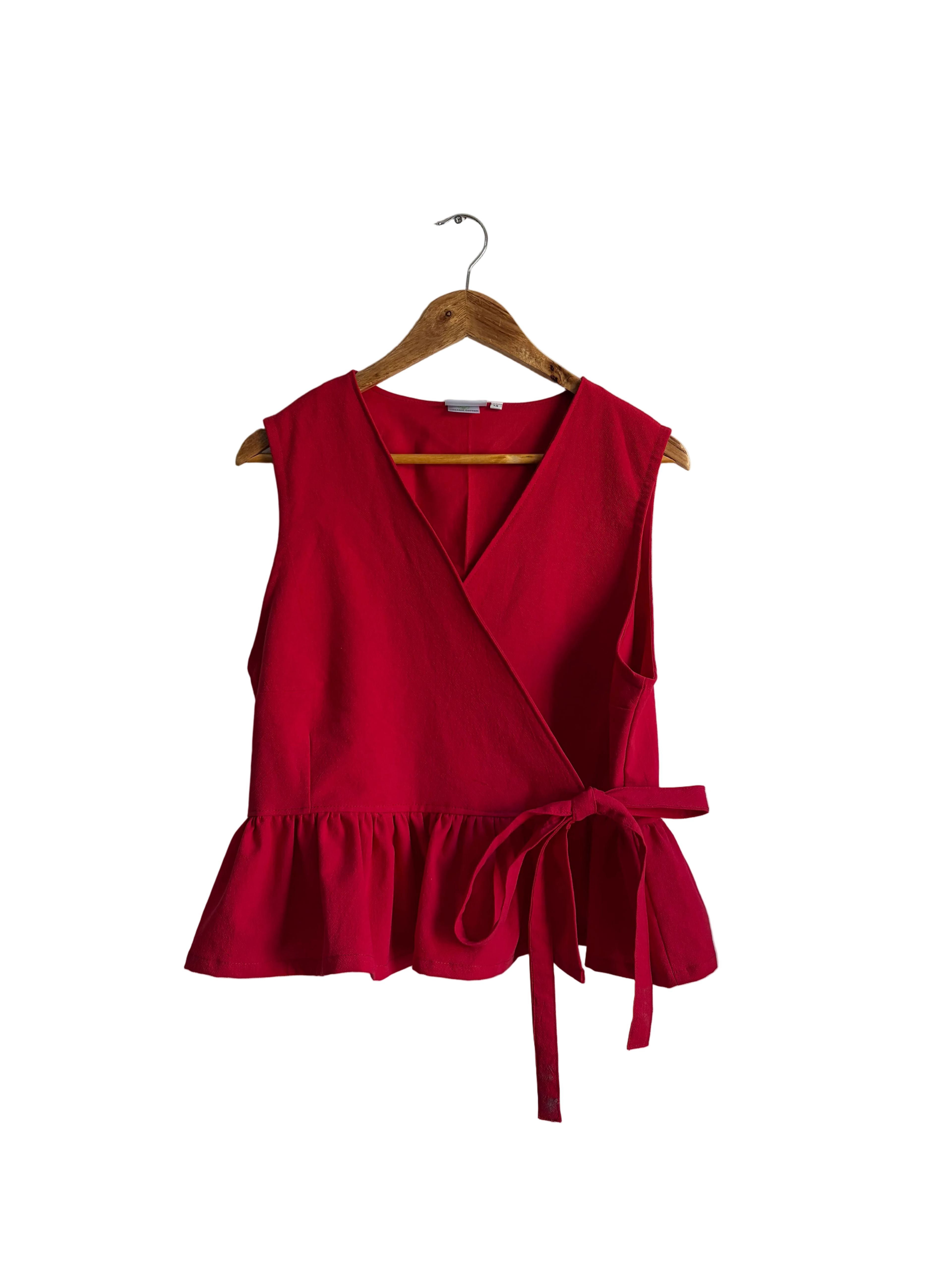 A bright red sleeveless wrap-style top with a ruffled hem and side tie, hanging on a wooden hanger against an off-white wall.