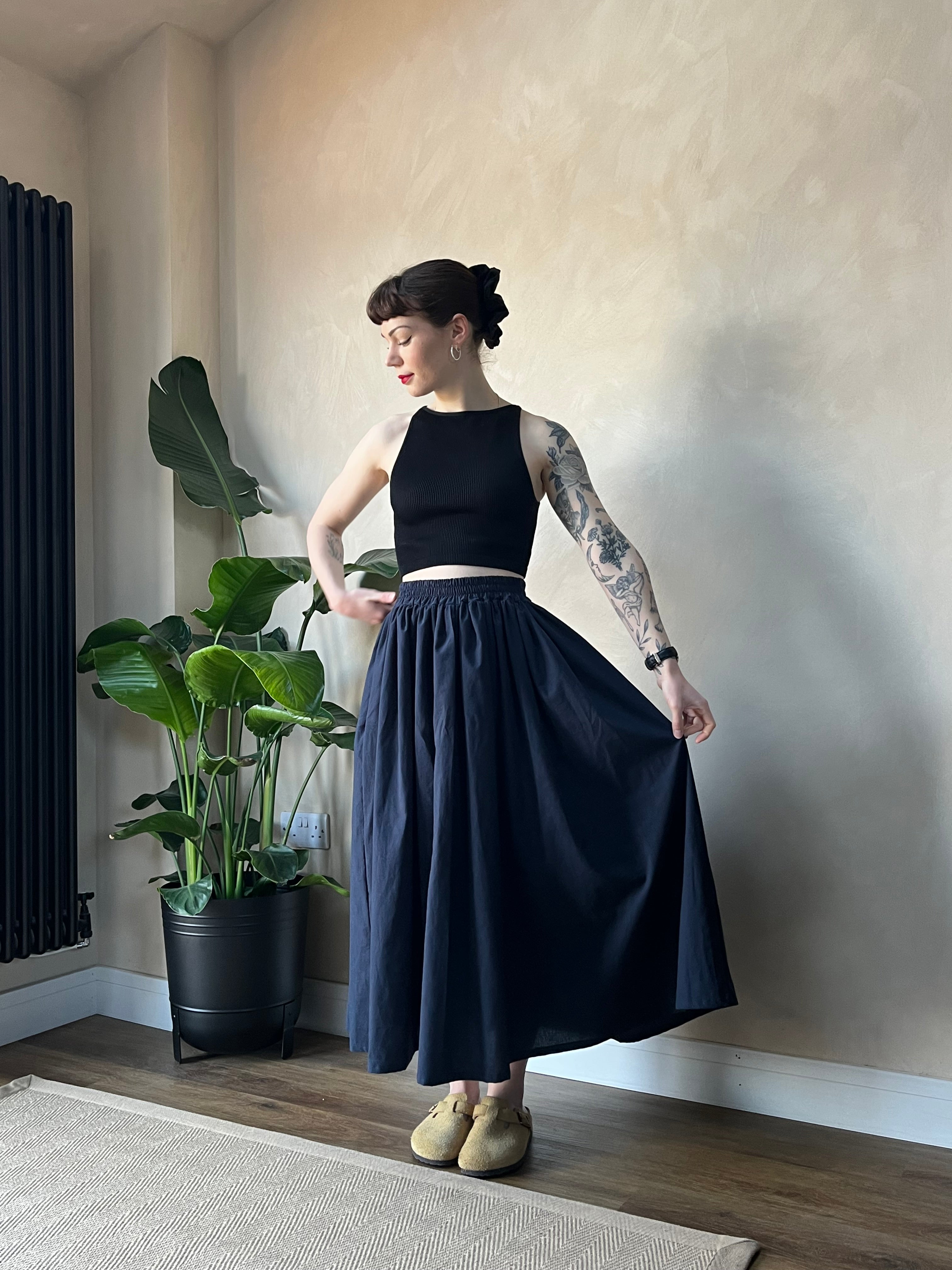 A hand holding a midi skirt in white, with a plant in the background. The skirt appears to be made from sustainable cotton fabric.