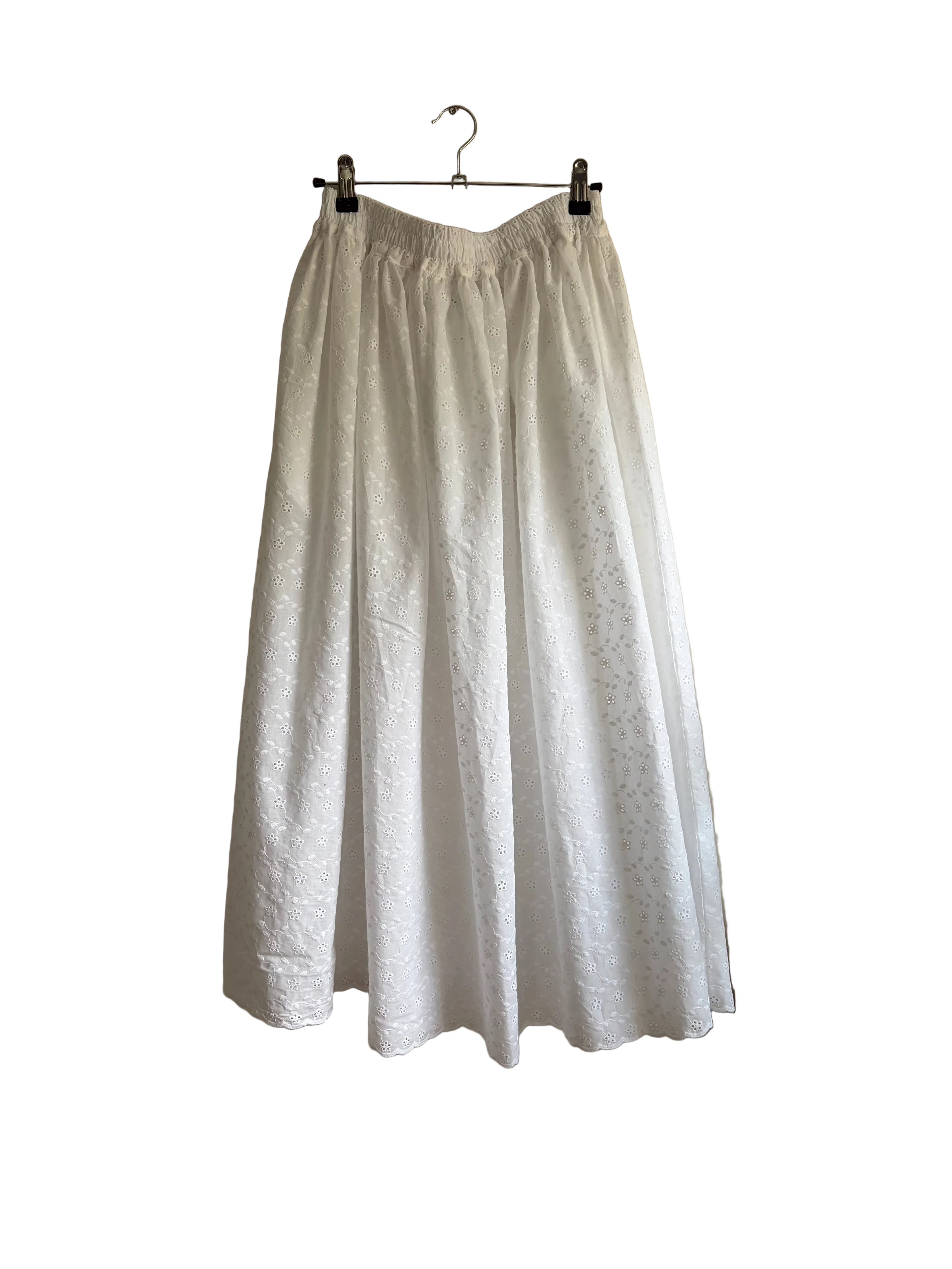A white textured midi skirt with an elastic waistband, hanging from a metal hanger against a beige wall next to a curtain.