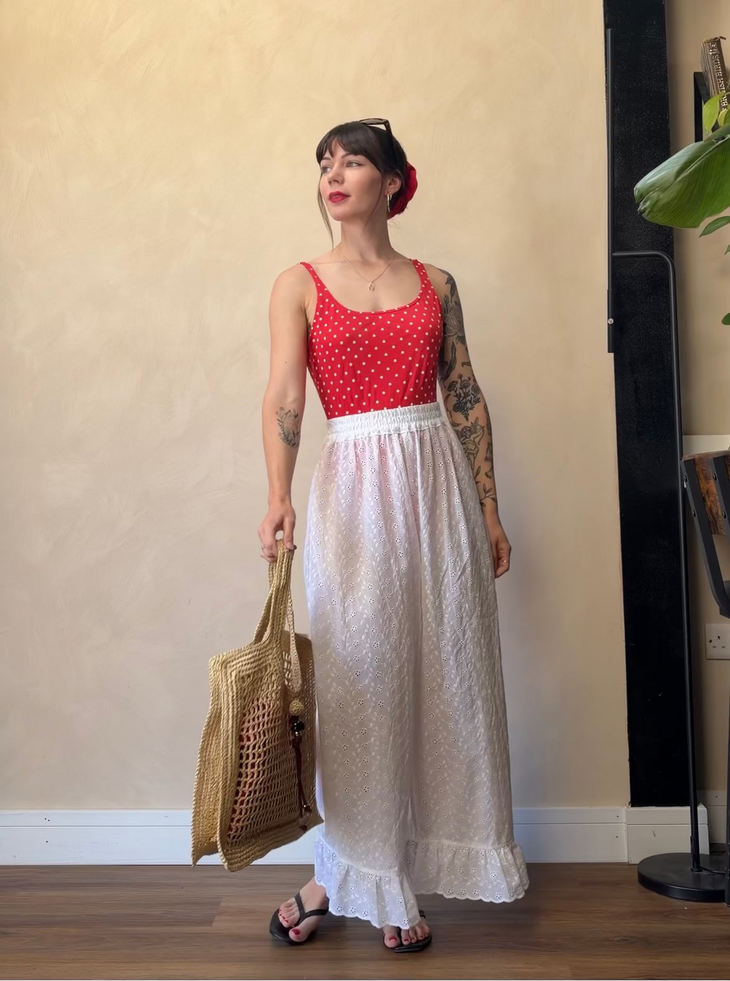 A woman stands indoors wearing a sleeveless red top with white polka dots and a long white skirt, holding a woven straw tote bag. She is posed against a beige wall on a wood floor, with a black floor lamp and a plant visible in the background.