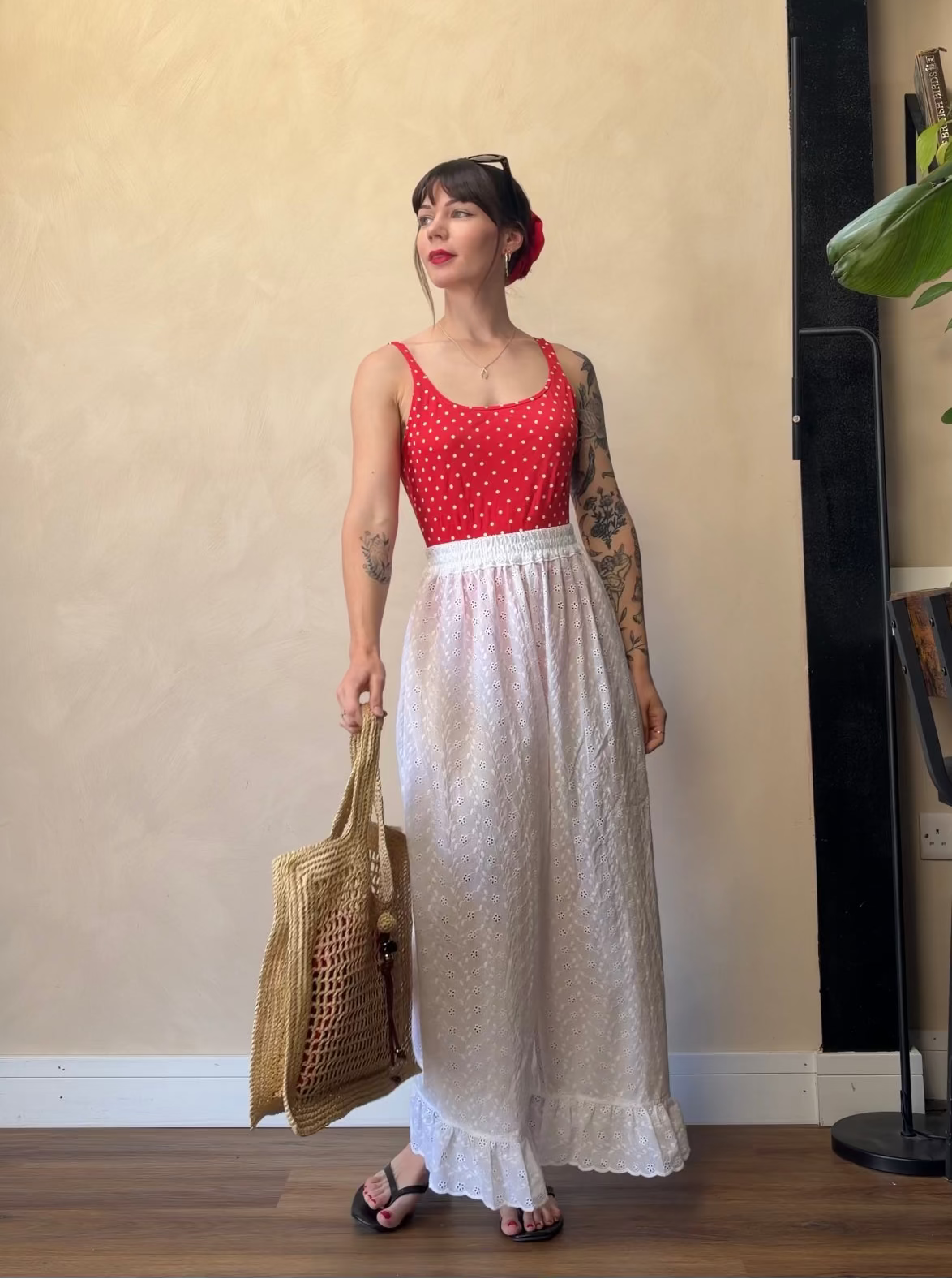 A woman stands indoors wearing a sleeveless red top with white polka dots and a long white skirt, holding a woven straw tote bag. She is posed against a beige wall on a wood floor, with a black floor lamp and a plant visible in the background.