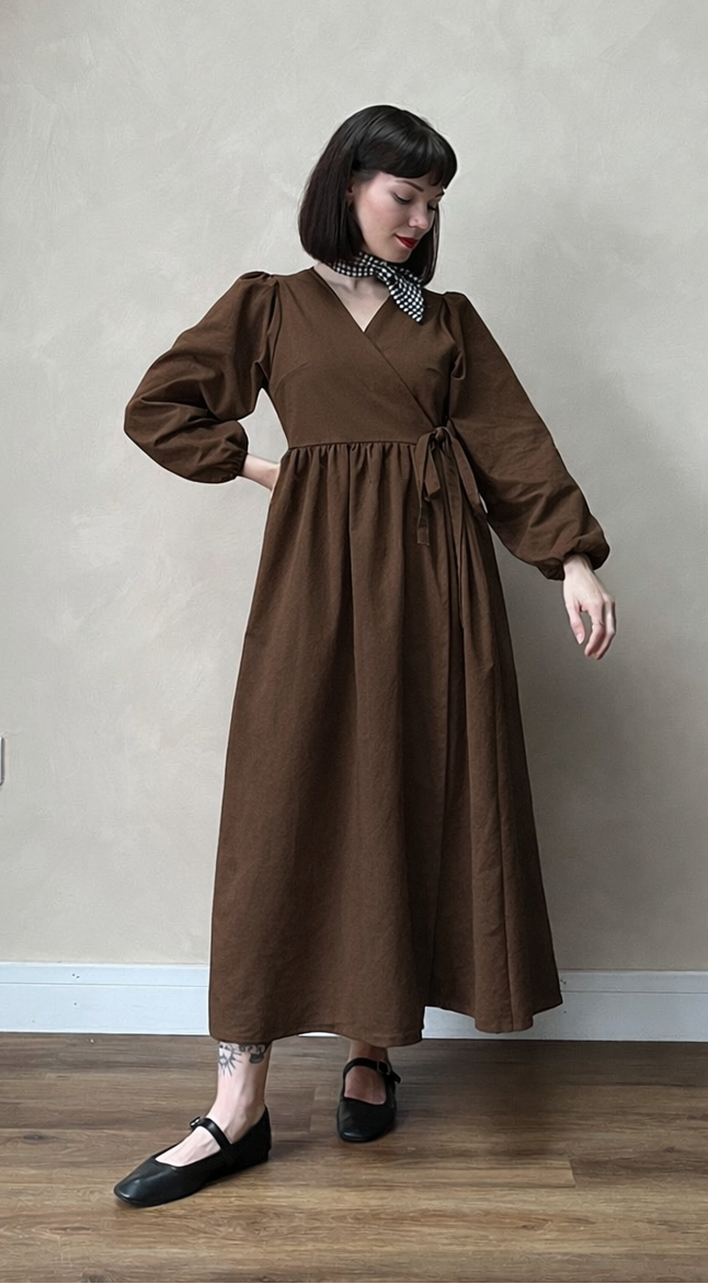Woman wearing a brown dress standing in a room with a neutral wall and wooden floor.